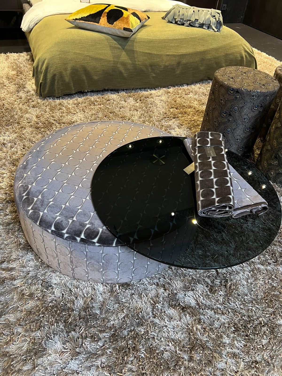 Runder Couchtisch mit Glasplatte, grauer Textilbezug, auf hellem Teppich, Bett im Hintergrund.