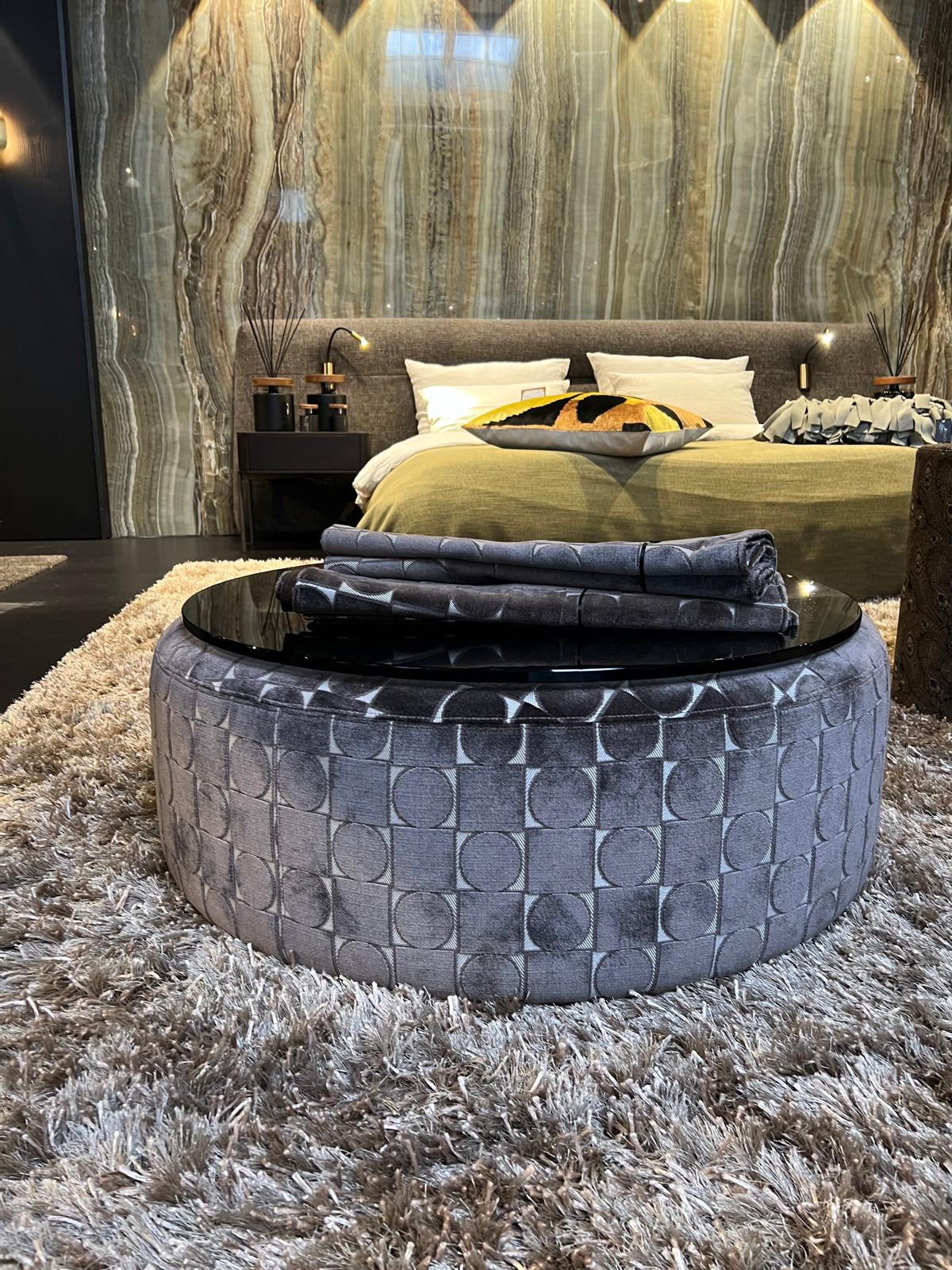 Grauer, gemusterter Sitzpouf vor einem Bett auf flauschigem Teppich in luxuriösem, modernem Schlafzimmer.