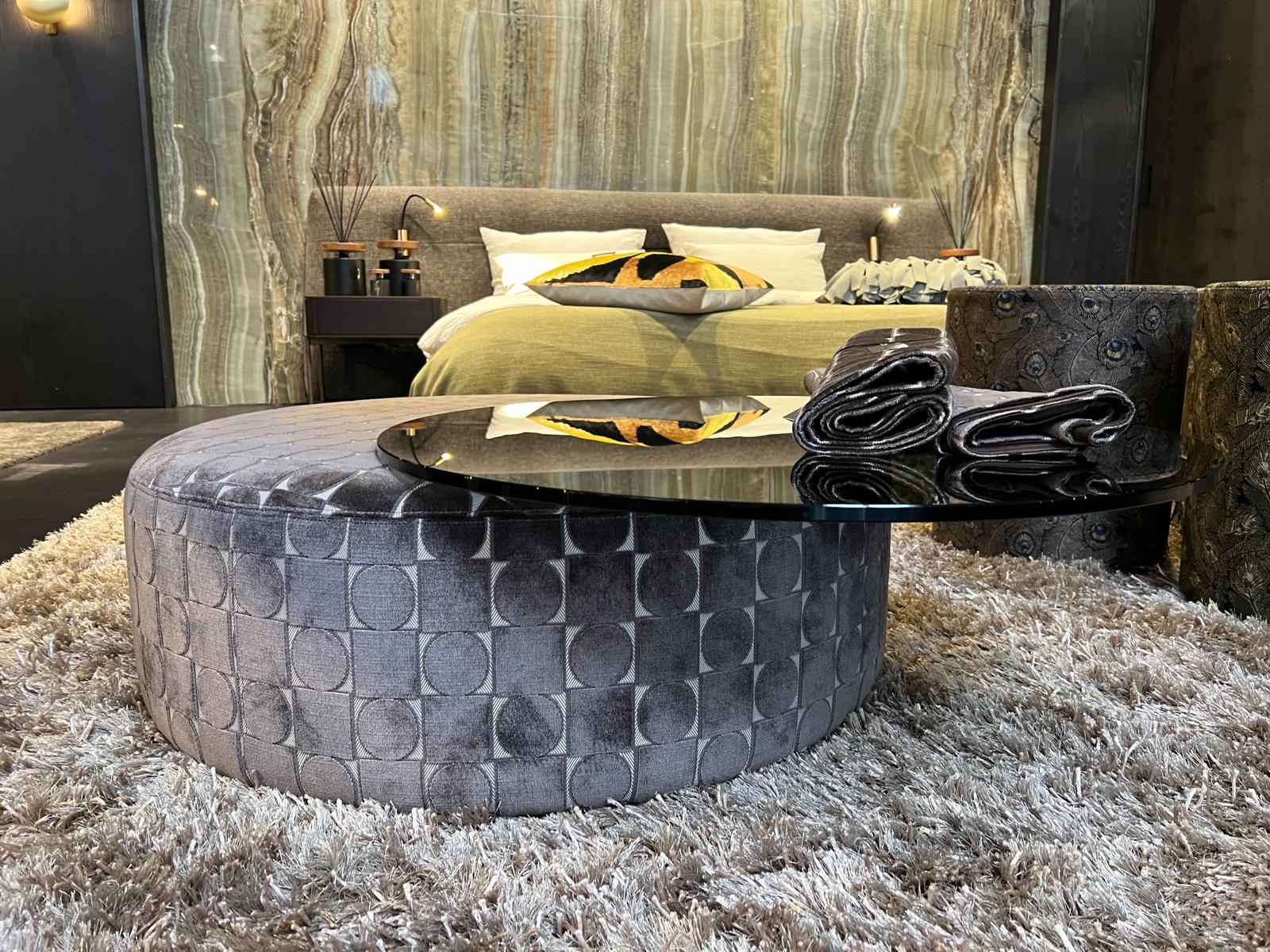 Gemütliches Schlafzimmer mit elegantem grauem Hocker und dekorativem Glastisch auf flauschigem Teppich.