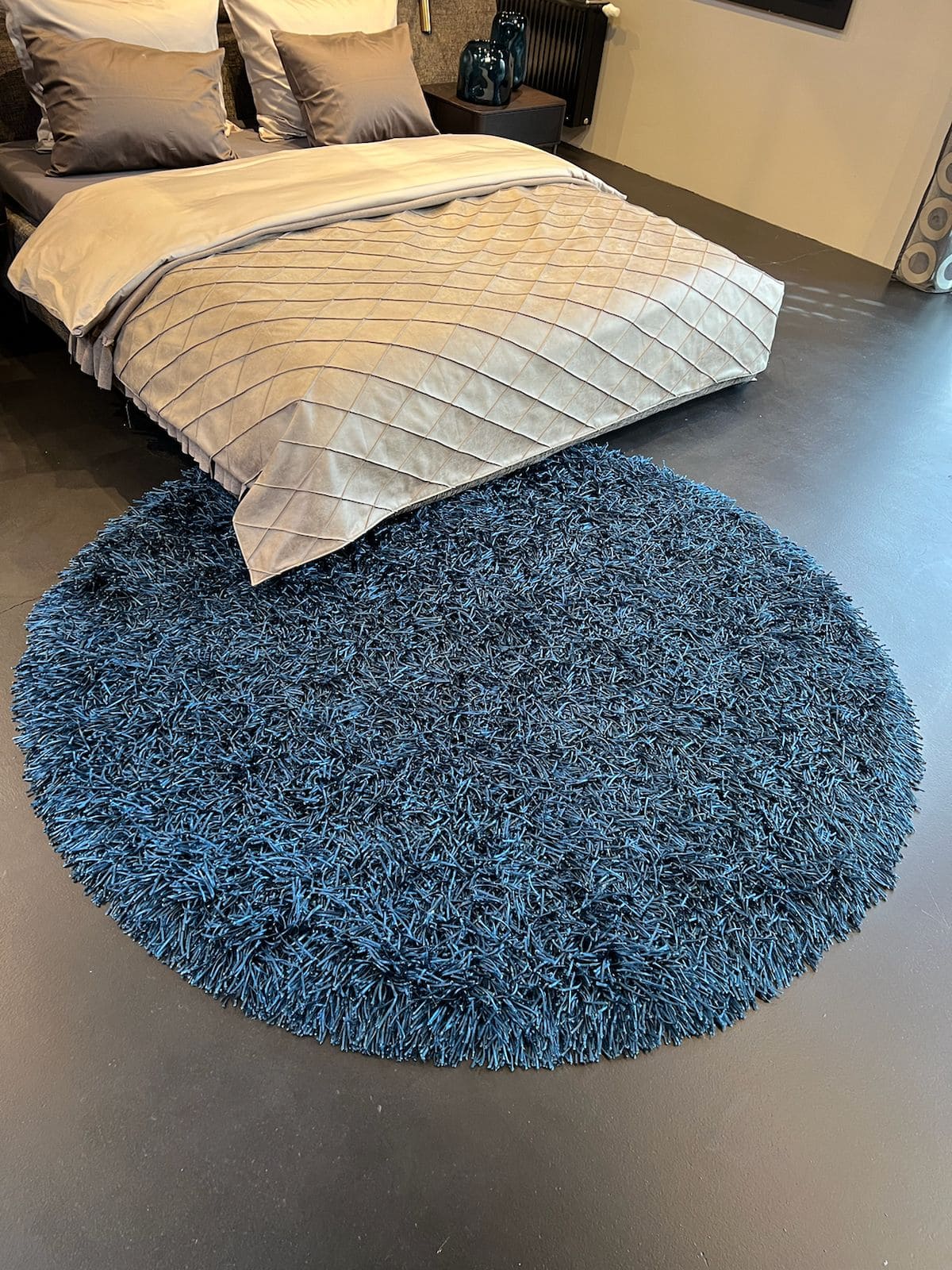 Runder blauer Hochflorteppich vor einem Bett mit beiger Tagesdecke